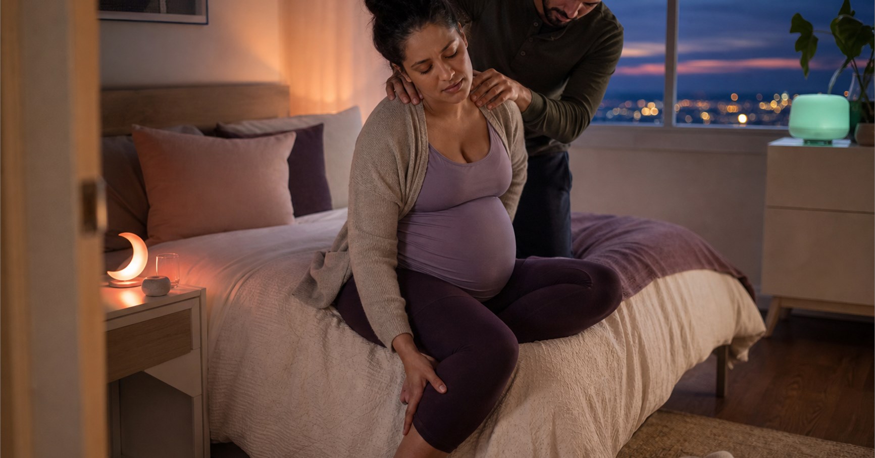 Pregnant woman sitting on a bed rubbing her calf while her partner massages her shoulders.
