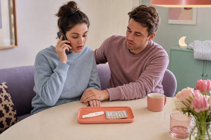 Woman calls a clinician while she and her partner look at a positive pregnancy test and a sealed birth-control pill pack on the table.