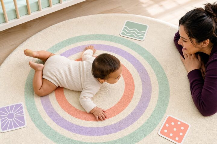 Awake baby pivoting on their tummy across a circular pastel play mat while caregiver watches nearby