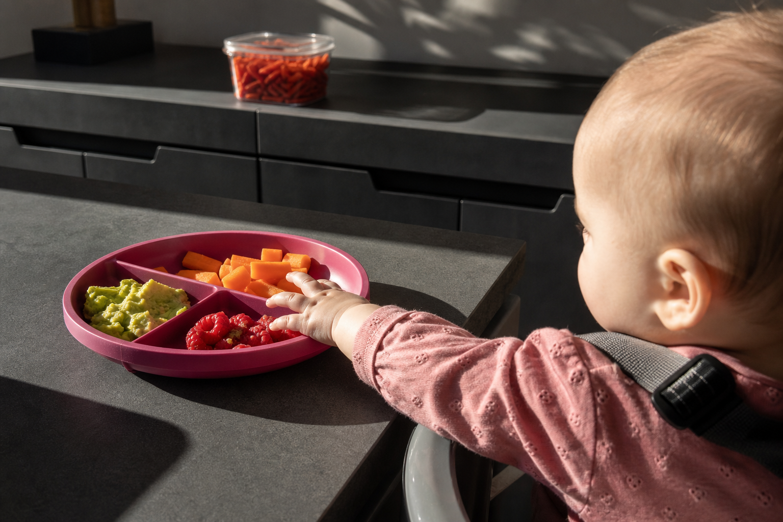 Baby reaches toward soft foods on a raspberry plate in a hotel kitchenette.