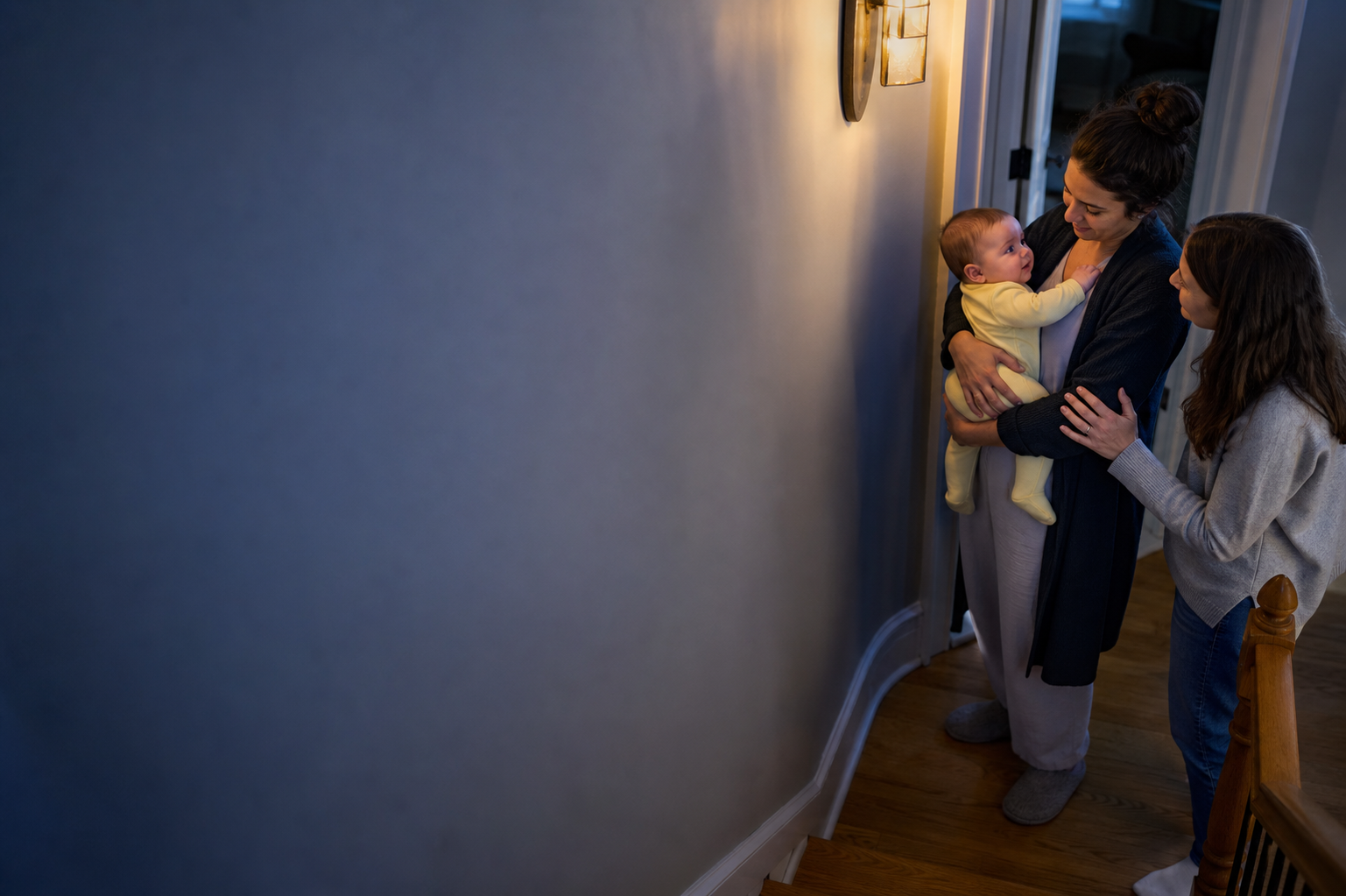 Caregiver holds an awake baby while a sleep coach offers reassurance in a blue-hour hallway.