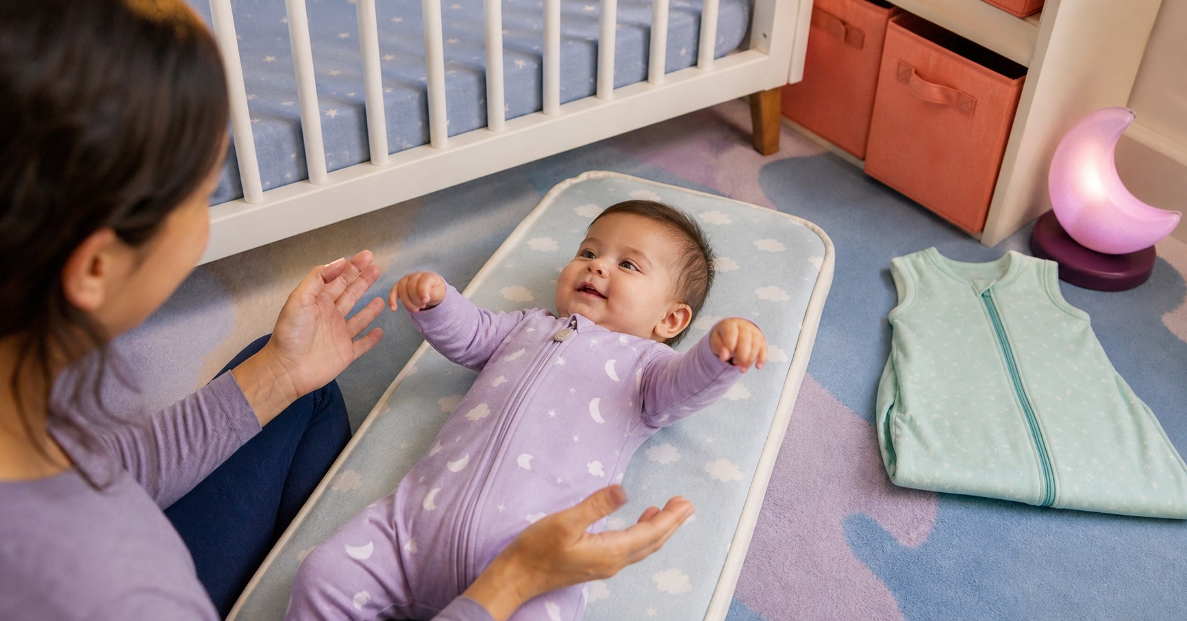 Smiling baby rotating both wrists on a floor changing mat while a parent watches calmly beside a bare crib