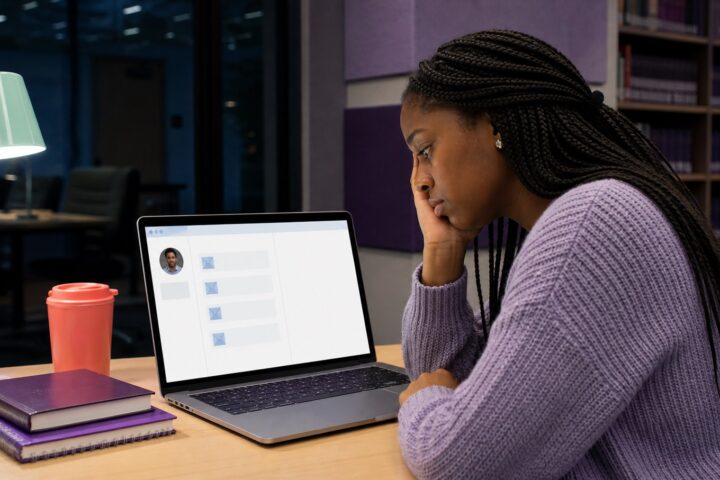 College student watches a laptop showing four sent emails and no reply while an empty faculty office sits behind her.