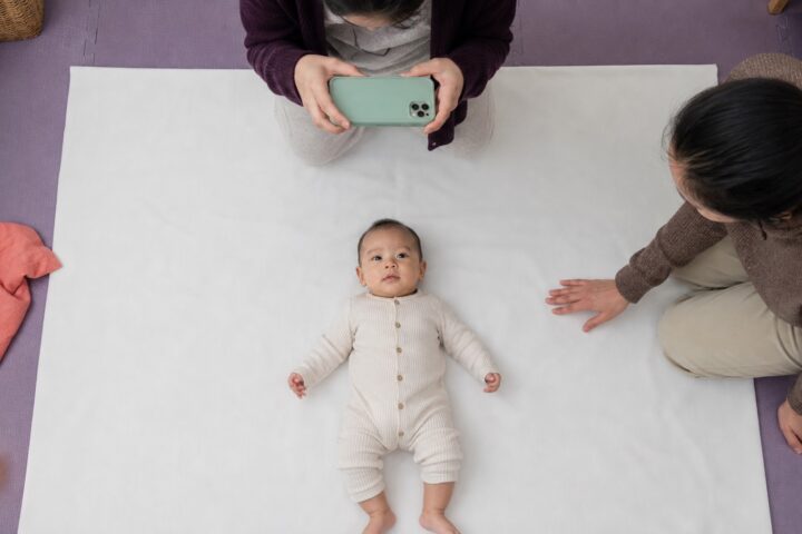 Awake baby lying safely on a plain white floor sheet while a caregiver takes an overhead phone photo