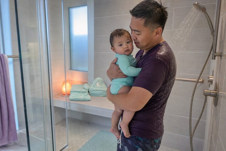 Father in a plum swim shirt holds his awake baby with both arms beside a running shower on a non-slip mat.