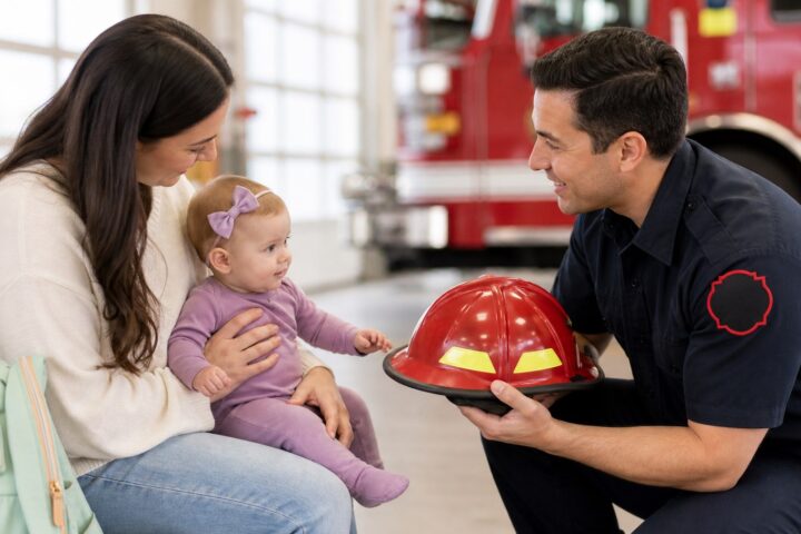A parent holds an awake baby as a firefighter crouches nearby and shows the baby a red helmet inside a fire station.