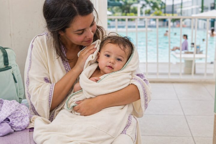 Mother drying her awake baby in a cream towel beside a fenced public pool