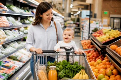 Caregiver pushing a grocery cart with a baby seated safely inside an aisle of produce and everyday items.
