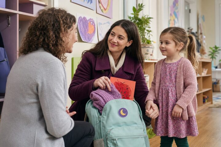 Parent packing a mint preschool backpack while holding a young child’s hand and talking with a teacher