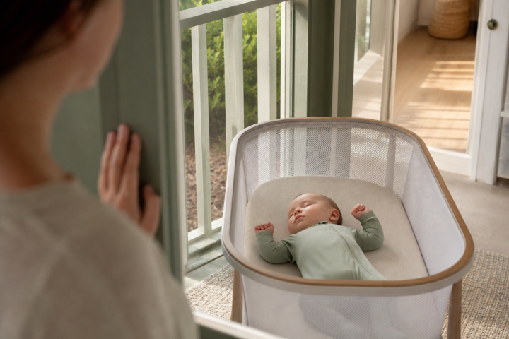 Baby sleeps on their back with raised hands in a porch bassinet as a caregiver watches.
