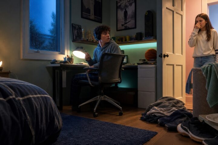 A teenage girl pinches her nose at a bedroom doorway as her teenage brother turns from his desk toward her.