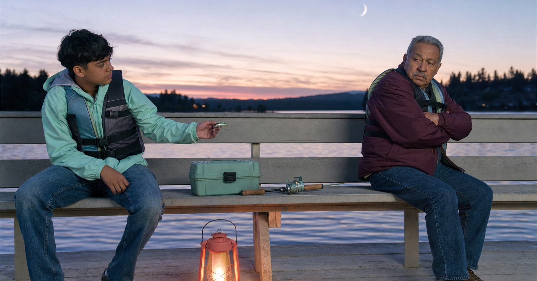 Teenage grandson offers a fishing lure to his guarded grandfather as they sit apart on a lakeside pier at dusk.