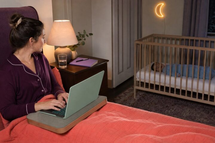 Parent working on a mint laptop from an adult bed while a baby sleeps safely in a separate bare crib
