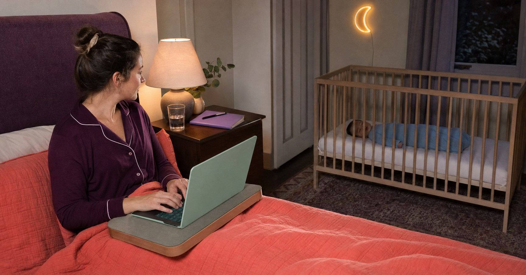 Parent working on a mint laptop from an adult bed while a baby sleeps safely in a separate bare crib