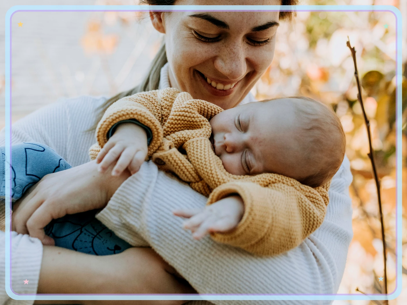 Smiling mother looks down at a sleeping baby held across her arms outdoors, framed by pale blue and lavender lines with tiny pastel stars