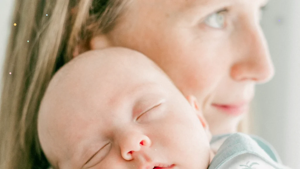 Awake caregiver's visible eye above a sleeping baby resting against the caregiver's shoulder, with small pastel points of light at the edges