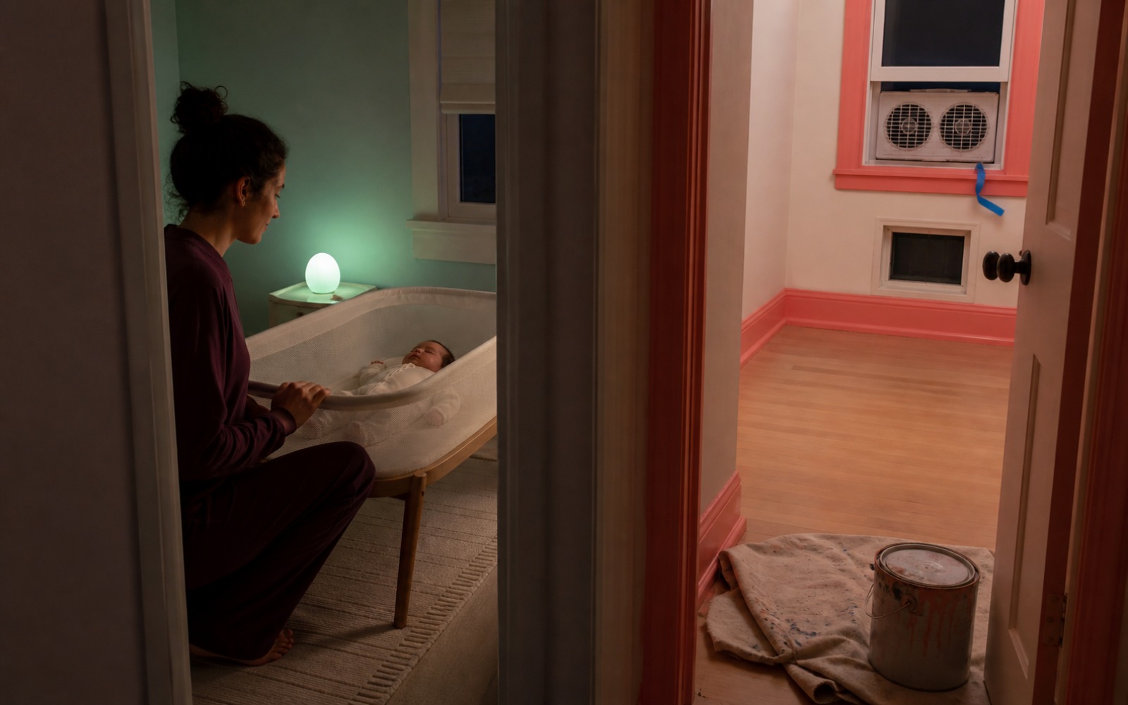 A clean alternate room holds a sleeping baby in a bare bassinet while a coral freshly painted nursery remains empty across the doorway