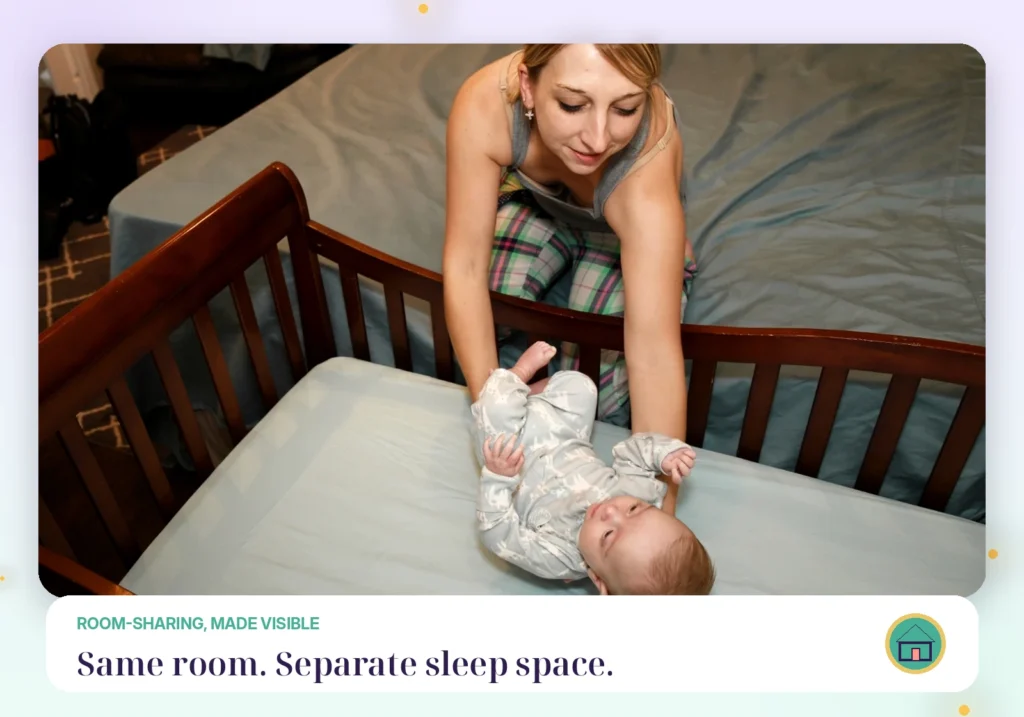 Mother reaching into a bare wooden crib beside an adult bed while a baby lies on their back on the fitted crib sheet