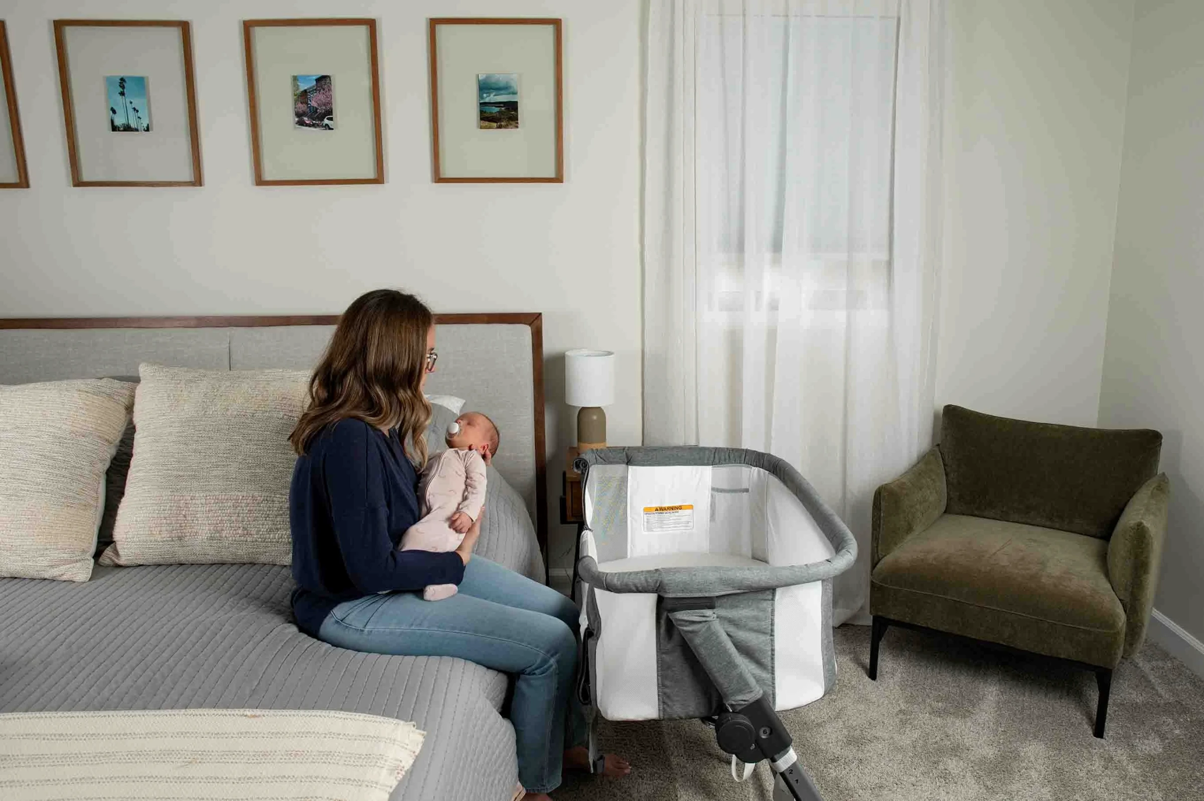 Mother sitting on a bed and holding an awake newborn beside an empty gray bassinet
