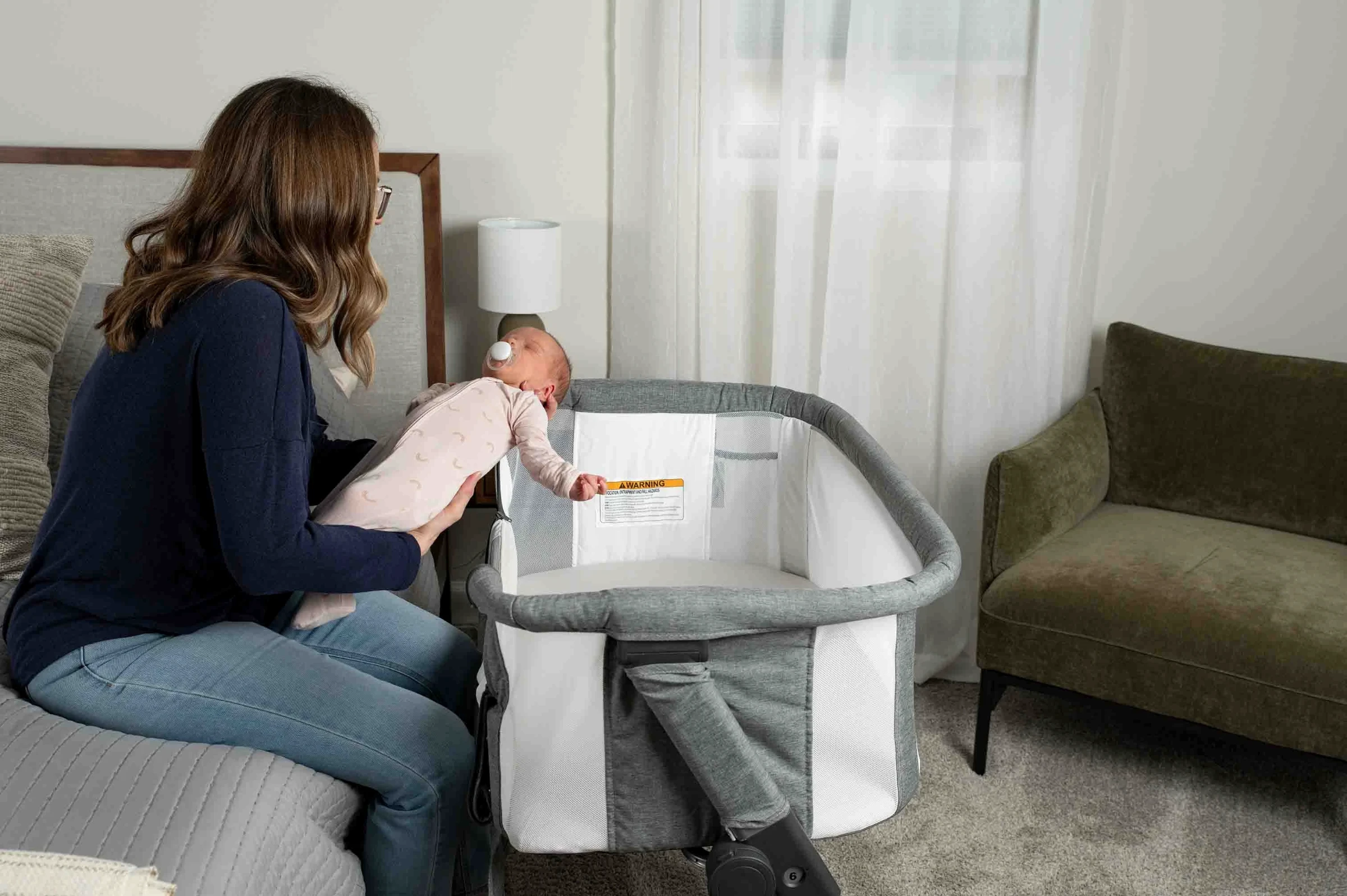 Mother supporting a newborn with both hands while lowering the baby toward an empty gray bassinet